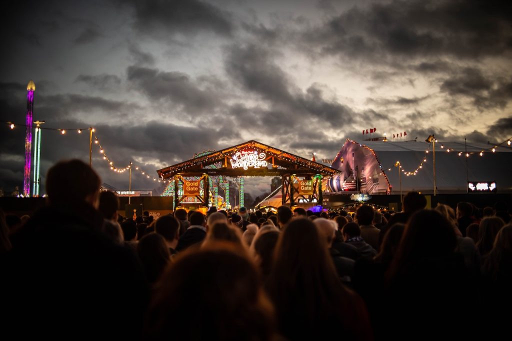 Centenas de pessoas de frente para um palco simples de madeira em um festival.